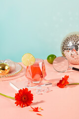 A glass containing a clear red liquid with a slice of lemon is placed on a glass podium, surrounded by fresh flowers and props. Pastel color background: pink and blue.