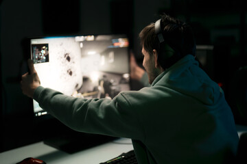 Back view of male cybersport gamer in headset touching computer monitor while celebrating successful game in dark room - obrazy, fototapety, plakaty