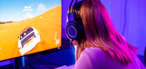 Happy Gamer endeavor plays online video games tournament with computer with neon lights, Young player woman wearing gaming headphones intend to do playing car games online at home, Back view - obrazy, fototapety, plakaty