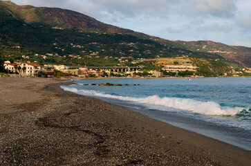 Picturesque autumn seascape the Tyrrhenian Sea near Patti, Sicily, Italy. Light waves and empty pebble beach. Cityscape in the background. Travel and tourism concept