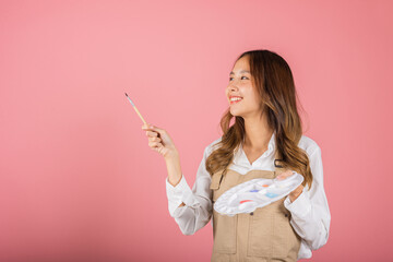 Asian beautiful young woman artist holding brush and paint palette, Happy female painting using paintbrush and palette with colors, studio shot isolated on pink background, Paintings and art equipment - obrazy, fototapety, plakaty