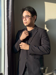 Vertical portrait of an Asian man with short, cool black hair, glasses, wearing a black business suit, buttoning the sleeves of his shirt. The background is the shadow of a window.