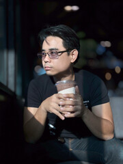 vertical portrait of an Asian man with short, cool black hair, wearing glasses, wearing a black shirt, is drinking coffee with a cafe in the background.