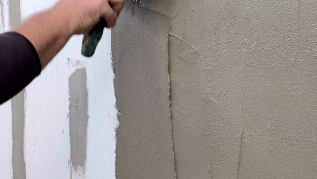 Hand with a putty knife, apply the putty to the wall. Process of applying a layer of putty. A man plasters the facade after insulating the building with polystyrene.