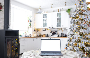 Laptop with a white screen mock up in the cozy white decorated Christmas kitchen with fairy lights and a Christmas tree. Seasonal remote work, internet, shopping, buying gifts Christmas and New Year.