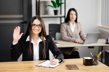Young happy businesswoman using notebook in modern office with colleagues. A girl in a business suit looking at the camera and making a hand gesture for greeting