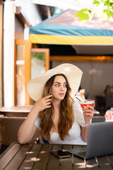 Beautiful stylish girl in a big hat drinking a cocktail and working as a freelancer sitting in a outdoors cafe