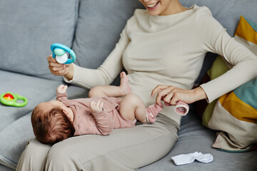Smiling woman sitting on couch and playing with newborn showing toy while taking baby socks off