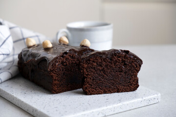 Chocolate pound cake with chocolate ganache on kitchen board with towel and cup of tea or coffee. Homemade bakery, classic recipe of chocolate loaf cake dessert.