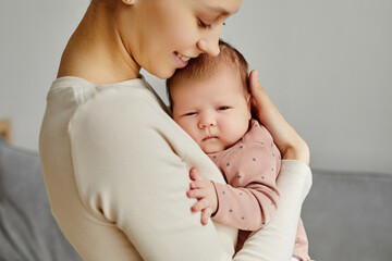 Side view at young smiling Caucasian woman hugging her sleepy newborn looking at camera, copy space