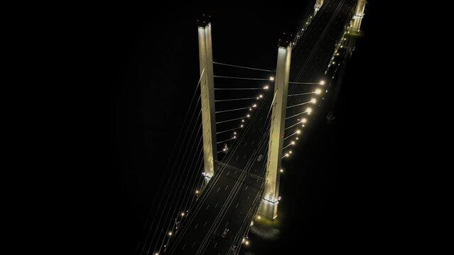 A cable-stayed highway bridge with tall concrete pylons illuminated by architectural lighting at night. General view from above from a drone