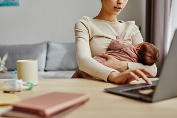 Close up of busy mother holding her baby while sitting at table and working on laptop at home, copy...