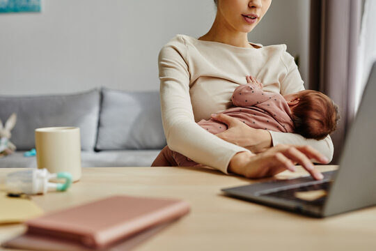 Close up of busy mother holding her baby while sitting at table and working on laptop at home, copy space
