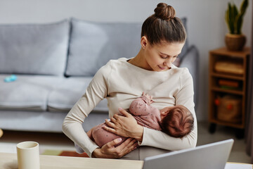 Waist up shot of happy Caucasian mother sitting by computer and lulling her baby during work break at home, copy space - obrazy, fototapety, plakaty