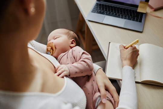 Top down view at sleeping newborn with pacifier in arms of mother working and writing down ideas, copy space