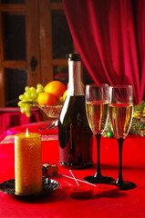A bottle of champagne, two glasses, a candle on a table with a red tablecloth.