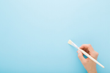Girl hand holding paintbrush on light blue wall background. Pastel color. Closeup. Empty place for...