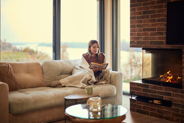 A woman reading a book while sitting on the sofa, covered with a blanket, enjoying herself alone. - obrazy, fototapety, plakaty