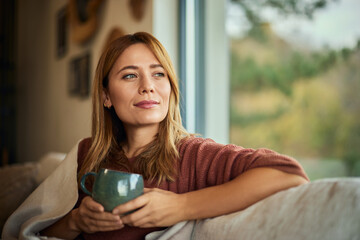A beautiful woman looking through the window, sitting on the sofa and enjoying a cup of tea.