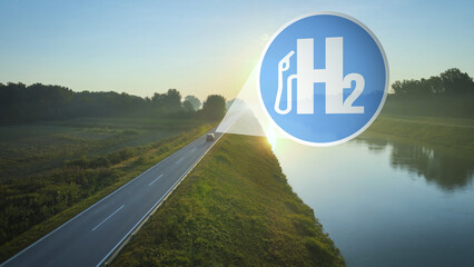 Hydrogen powered car drives into the sunset on an idyllic road at edge of river. H2 Sybol Concept...