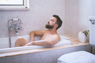 Bearded man enjoy and relaxes in bath time in beige vintage bathroom. Skin and self-care concept,...
