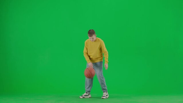 Portrait of boy on chroma key green screen. School boy kid in casual clothing playing with basketball ball, swings it on finger.