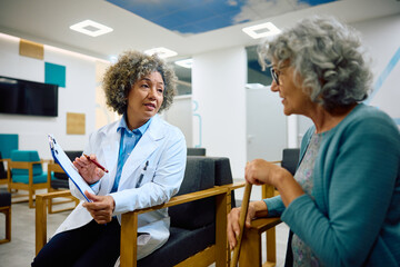 Female doctor analyzing senior woman's medical data while communicating with her in waiting room at clinic.