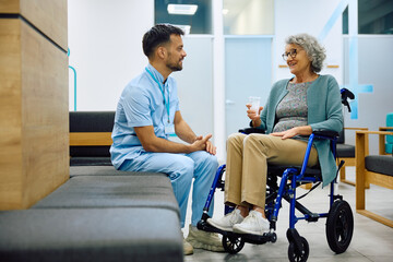 Happy senior woman in wheelchair talks to medical technician in hallway at clinic. - obrazy, fototapety, plakaty