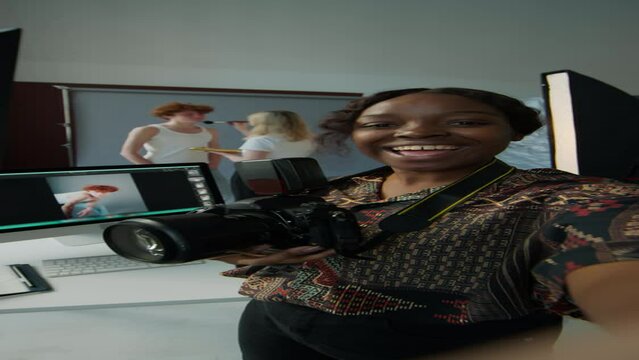 Vertical POV shot of young Black female photographer holding digital camera and telling about preparation for fashion photo shoot in studio, having video call or live streaming