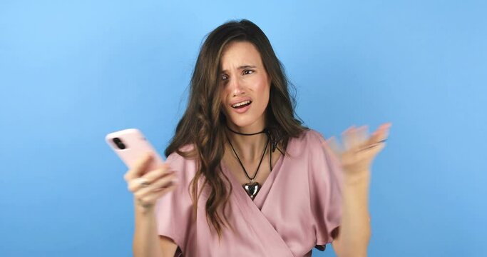 Mad and shock young brunette beautiful woman using mobile phone isolated on blue background. Yelling unhappy woman hold mobile phone. Disappointed sad upset lady horrified impressed news. WTF. Oh no