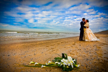 The image depicts a poignant moment between a bride and groom, embracing on a sandy beach with the...