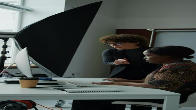 Vertical shot of female stylist and photographer looking at reference picture on computer screen and having discussion while preparing for shoot in studio