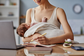 Cropped shot of mother writing down ideas in front of computer and holding her baby in white...