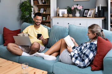 Happy young man working on sofa with laptop near woman with book lying on sofa in living room at...