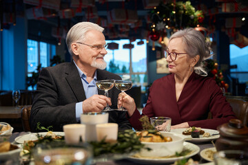 Joyful aged man and woman celebrating Christmas together clinking glasses