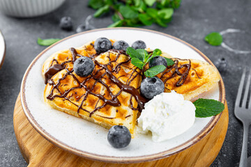 Homemade waffles with blueberries, chocolate sauce and cream cheese in a plate and a grey background close up