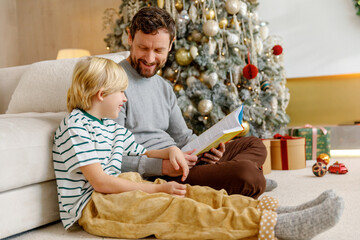 father and little son sit on floor near christmas tree at home, and read fairy tale book together, smile and laugh at funny stories