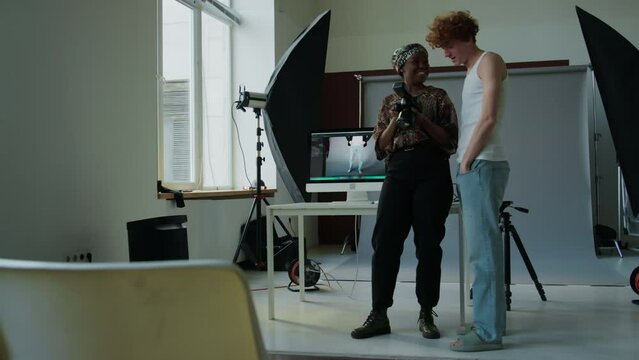 Full length shot of young male fashion model and African American female photographer checking and discussing pictures on camera after studio shoot
