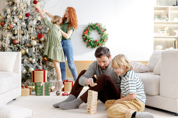 dad and son play jenga together on floor at home, smiling and laughing, mom and daughter decorate...