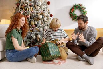 Obraz na płótnie Canvas mom, dad and son sit together on floor in living room near decorated christmas tree, boy unwrap his present. feel happy, joyful and inspired, smiling and laughing