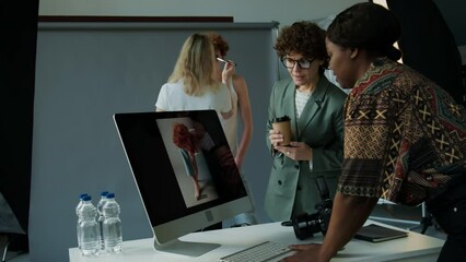 Female creative director holding coffee cup and discussing image on computer screen with photographer during shoot day in studio, stylist preparing model in backgroundFemale creative director holding 