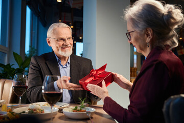 Smiling aged man receives present box from woman during New Year Eve celebration