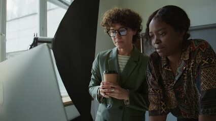 Multi-ethnic female art director and photographer checking images on PC and discussing results of shoot day in studio