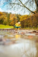 Sun always shines after the rain. Small bond infant boy wearing yellow rubber boots and yellow waterproof raincoat walking in puddles in city park on sunny rainy day