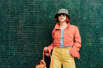 Hipster young woman in trendy peach color jacket, sun glasses, backpack bag and bucket hat posing...