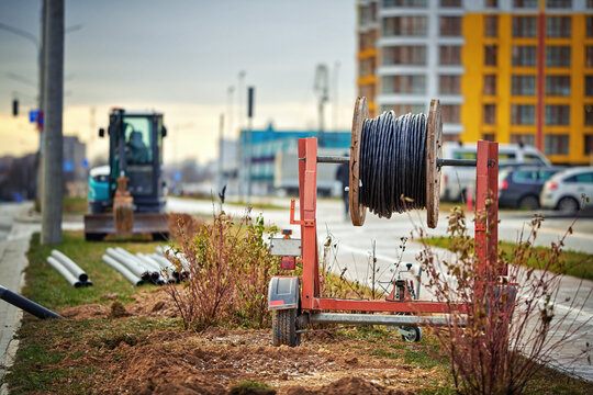 Wooden drum with fiber optic cable mounted on trailer, excavator digging trench for cable on background. Laying optical fiber cable in residential district. Underground communications.
