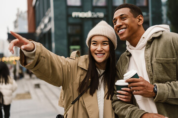 Cheerful couple pointing away while drinking coffee standing at street
