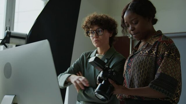 Young diverse female photographer and creative director checking photos on camera and computer and discussing shoot day in studio