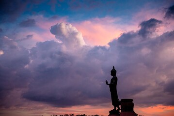 Stand big Buddha Statue in silhouette sun set Light background in park of Thailand temple.Yellow orange light silhouette dark shadow of image Buddha statue stand Buddha tall walk in sun light cloud. - obrazy, fototapety, plakaty