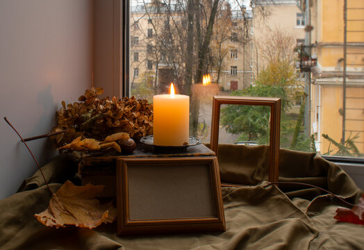 Empty wooden frames for photos or paintings, a burning candle on a stack of old books and dry autumn leaves on the windowsill against the backdrop of a window with an urban landscape.